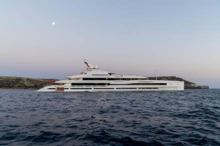 A luxurious white superyacht glides through the calm, dark blue ocean under a twilight sky with a visible moon.