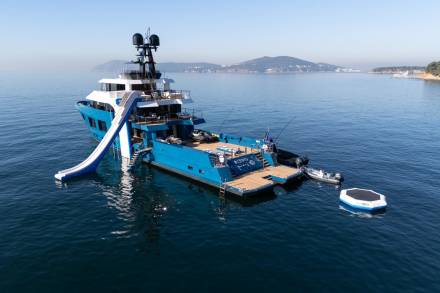 A vibrant blue yacht with a water slide and inflatable island awaits adventure on the tranquil sea.