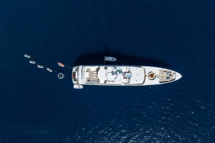 An aerial view reveals a sleek white yacht anchored in deep blue waters, surrounded by several smaller boats and inflatable toys.