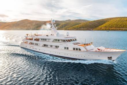 A majestic white yacht cruises through sparkling blue waters against a backdrop of sun-drenched hills.