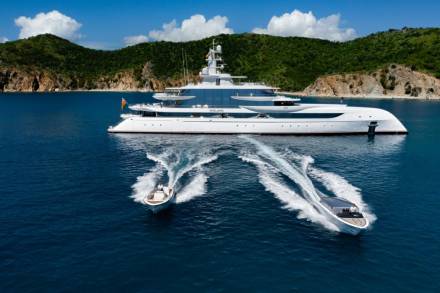 A sleek white superyacht dwarfs two speedboats leaving white wakes in the deep blue ocean near a lush green island.