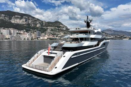 A luxurious superyacht glides through calm blue waters with a picturesque coastal city nestled against a mountainous backdrop.