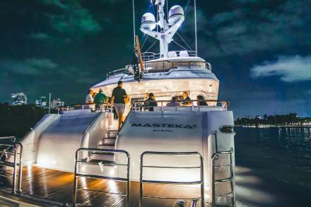 A luxurious yacht cruises through dark waters at night, its deck illuminated by warm lights and a group of people enjoying the evening.