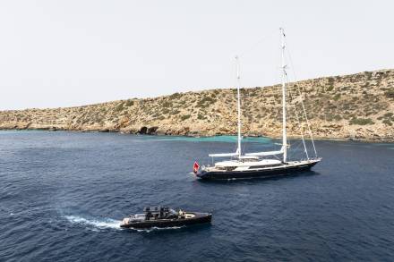 A sleek black tender speeds through the deep blue water near a majestic sailing yacht anchored off a rocky, sun-drenched island.