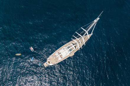 An aerial view captures a grand sailing yacht surrounded by smaller boats and water toys on a deep blue sea.