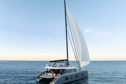 A sleek, modern catamaran yacht glides across the calm ocean under a clear blue sky with its sails billowing in the breeze.