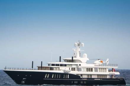 A sleek, dark-hulled superyacht with a helipad glides across choppy blue waters under a vast sky.