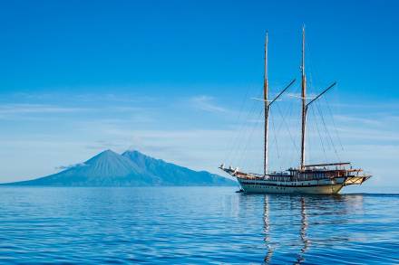 A majestic schooner glides across calm blue waters with a volcanic island silhouetted against a clear sky.
