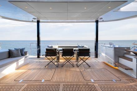 A spacious outdoor deck with a panoramic view of the ocean, featuring a seating area with chairs and a table, surrounded by a glass enclosure that provides shelter from the elements.