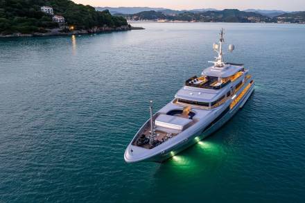 A large, luxurious yacht is anchored in a serene, turquoise bay surrounded by lush, verdant hills and buildings in the distance.