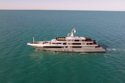 A large, luxurious yacht is sailing across a calm, turquoise sea under a clear, pastel-colored sky.