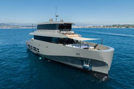 A sleek, modern gray yacht with an open deck area is anchored in calm blue water, with a distant coastline and clear sky in the background.
