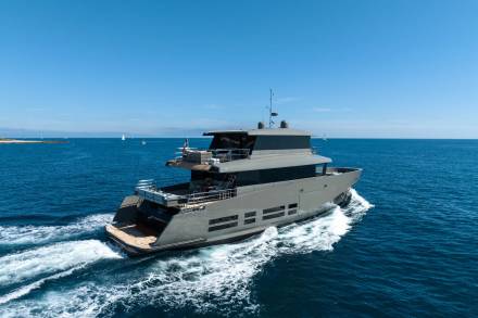 A sleek, modern gray yacht cruises through calm blue waters, leaving a white wake, with a distant coastline and a clear sky in the background.