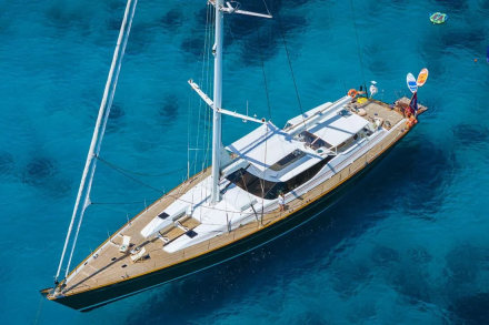 A large white sailboat with a wooden deck floats on crystal clear turquoise water, with a few colorful inflatable toys visible in the distance.