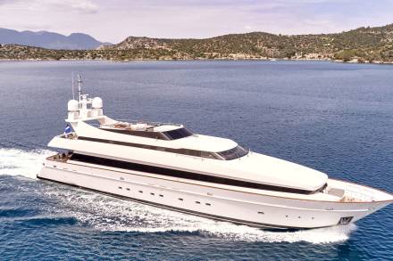 A sleek white yacht cruises through calm blue waters, with a verdant, hilly coastline dotted with buildings in the background.