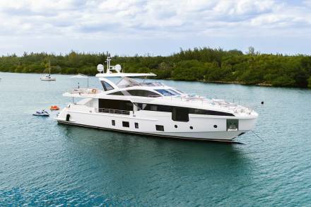 A sleek white yacht floats on calm blue water in the foreground, with lush green trees and a cloudy sky forming the background.