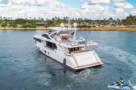 A luxurious white yacht cruises on calm blue water in the foreground, with a jet ski trailing behind, while palm trees and a distant cityscape line the background.