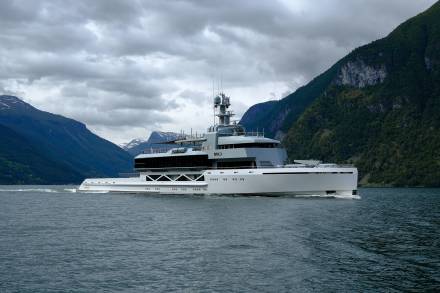 A sleek, modern yacht cruises through calm water with dramatic, cloud-covered mountains rising in the background.