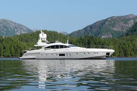 A sleek white yacht floats on calm water in the foreground, with a lush green forest and mountains filling the background.