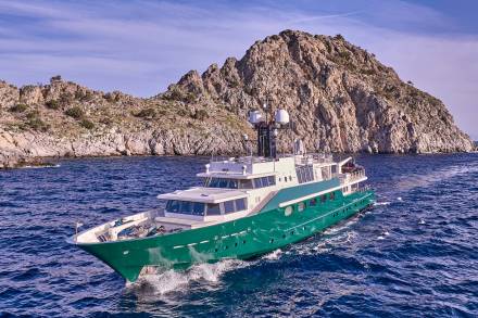 A green yacht cruises through blue water in the foreground, with a rocky, sunlit island in the background under a clear sky.