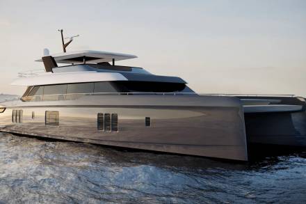 A sleek, modern catamaran yacht glides across calm, rippling water under a soft, overcast sky.
