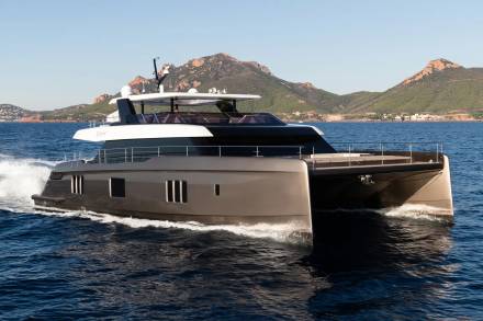 A sleek, modern catamaran yacht cruises through calm blue water with a rugged, mountainous coastline in the background.