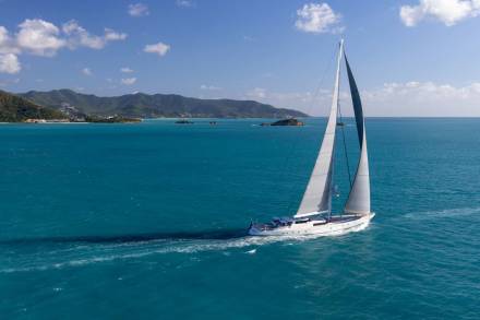 A white sailboat with full sails glides across turquoise water, leaving a foamy wake, with a lush green coastline and distant islands under a bright blue sky in the background.