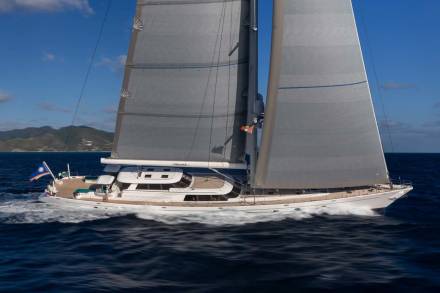 A large white sailboat with full sails glides through dark blue water, with green, hilly islands visible in the hazy background under a bright blue sky.