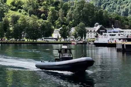 A dark inflatable boat speeds across calm water in the foreground, with a village nestled at the base of a lush, green mountain in the background.