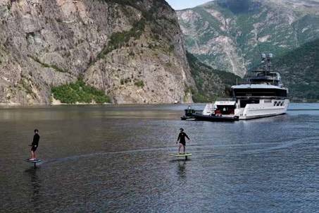 Two people on hydrofoils glide across a calm lake in the foreground, with a large white yacht anchored in the background against a backdrop of steep, tree-covered mountains.