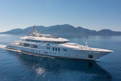 A sleek white superyacht glides through calm blue waters with a mountainous coastline in the background.