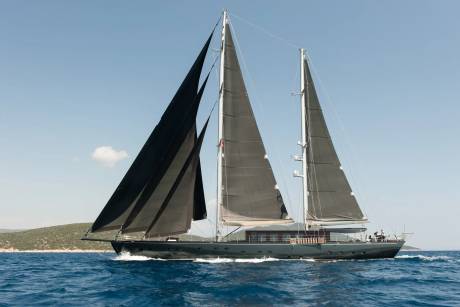 A sleek, dark-sailed yacht cuts through the vibrant blue ocean with a lush green coastline in the background.