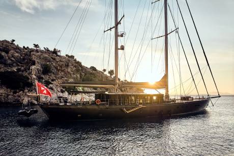 A majestic yacht glides through calm waters near a rocky, tree-dotted coastline as the sun sets.