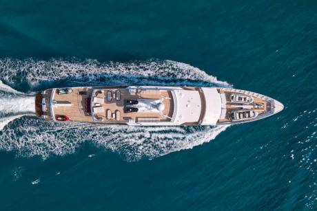 An aerial view captures a luxurious white yacht cruising through the sparkling blue ocean, leaving a foamy wake behind it.