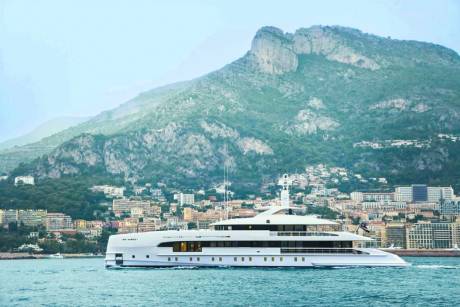 A large white yacht sails on the water in the foreground, with a densely populated coastal city and a rugged mountain in the background.