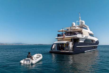 A man in a small dinghy approaches a large, luxurious yacht anchored in calm blue water with a distant coastline under a clear sky.