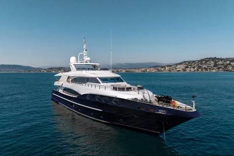 A luxurious dark blue and white yacht floats on calm blue water in the foreground, with a lush green, hilly coastline under a clear blue sky in the background.