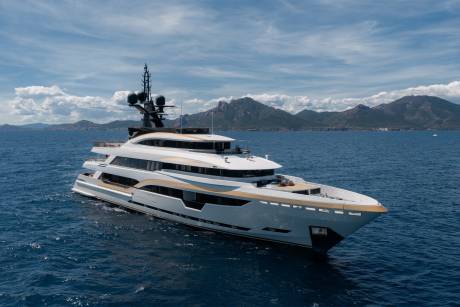 A luxurious white and gold yacht cruises on calm blue water with a mountainous coastline under a cloudy sky in the background.