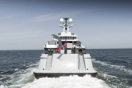 A large white yacht with a red flag sails through calm blue water, leaving a foamy wake under a clear sky.