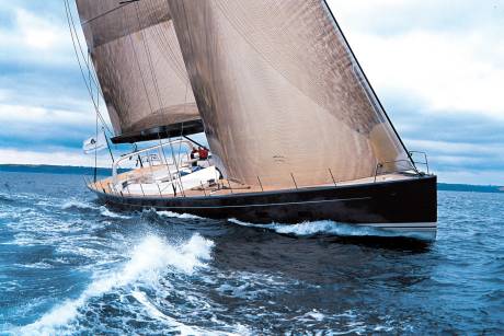 A sleek, dark sailboat with billowing sails cuts through choppy blue water, with a hazy coastline visible in the distance under an overcast sky.