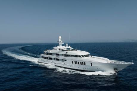 A large white yacht cruises through the deep blue ocean, leaving a foamy wake against a clear sky.