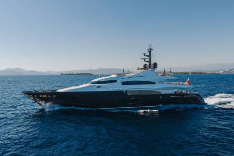A sleek, dark-hulled luxury yacht cruises through deep blue waters against a backdrop of distant mountains and a clear sky.