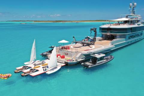 A luxurious superyacht is anchored in crystal-clear turquoise waters, surrounded by a fleet of watercraft and toys.