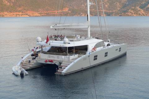 A large white catamaran anchors in calm waters with a mountainous coastline in the background.