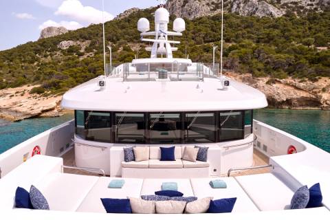 Luxurious white yacht decks are adorned with plush cushions and towels, set against a backdrop of lush green hills and sparkling turquoise waters.
