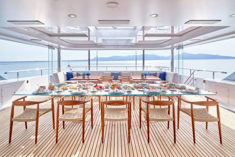 A beautifully set dining table awaits on a yacht's deck, offering a scenic view of the sea and distant mountains.