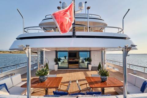 A large yacht with a red flag flying proudly is docked at sea, featuring a spacious deck with seating and dining areas.