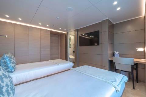 This modern yacht cabin features two twin beds, a wall-mounted television, and a sleek desk area with a chair.