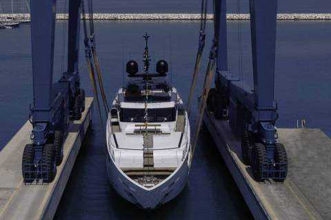 A sleek white yacht is lifted from the water by a large blue boat lift in a sunny marina.
