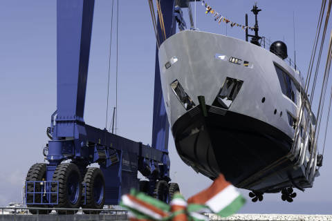 A massive blue straddle carrier lifts a sleek white yacht against a clear blue sky.